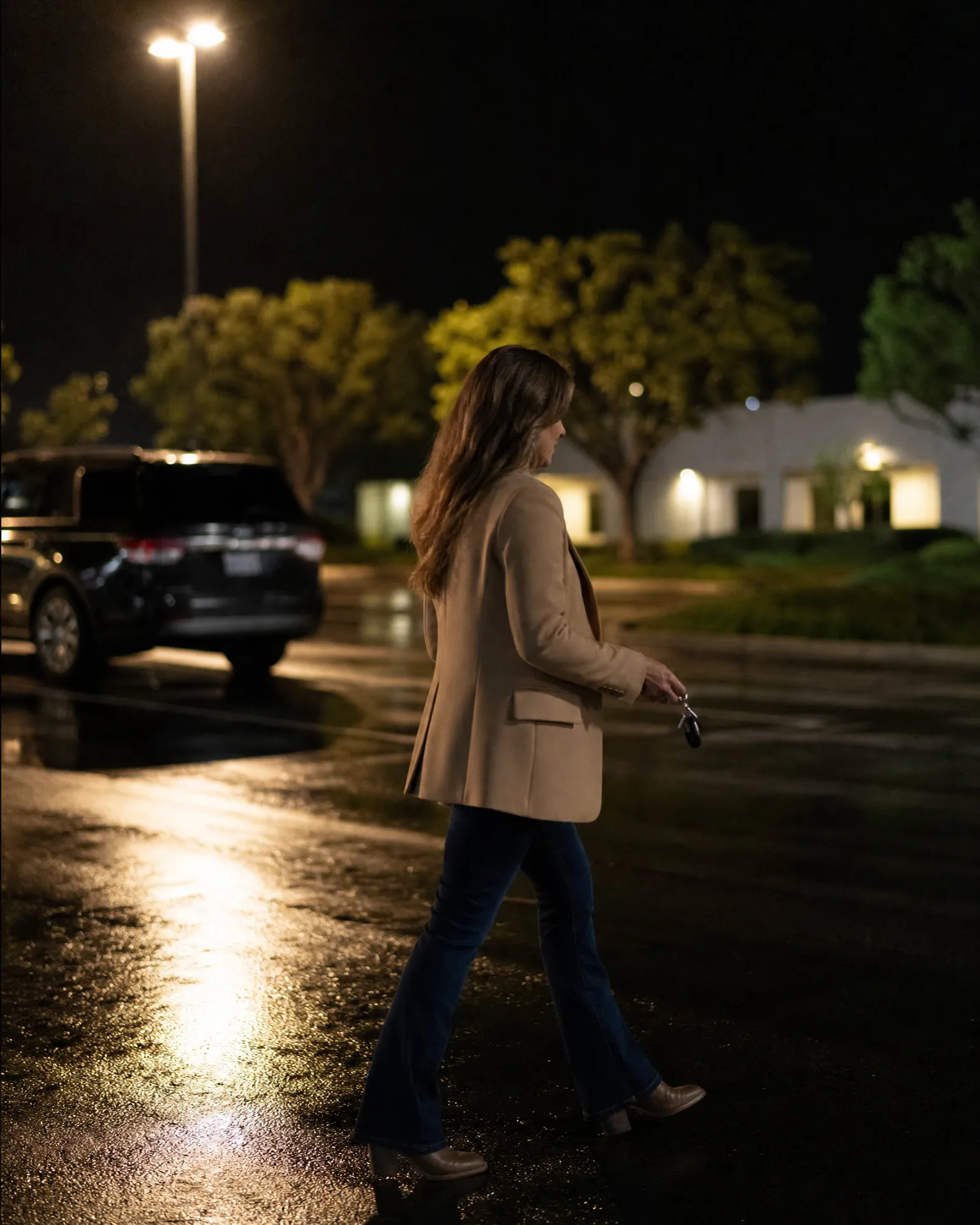 A woman in a tan blazer, jeans and heeled boots walks across a wet parking lot at night, holding keys, with an SUV, trees and a low white building lit behind her.