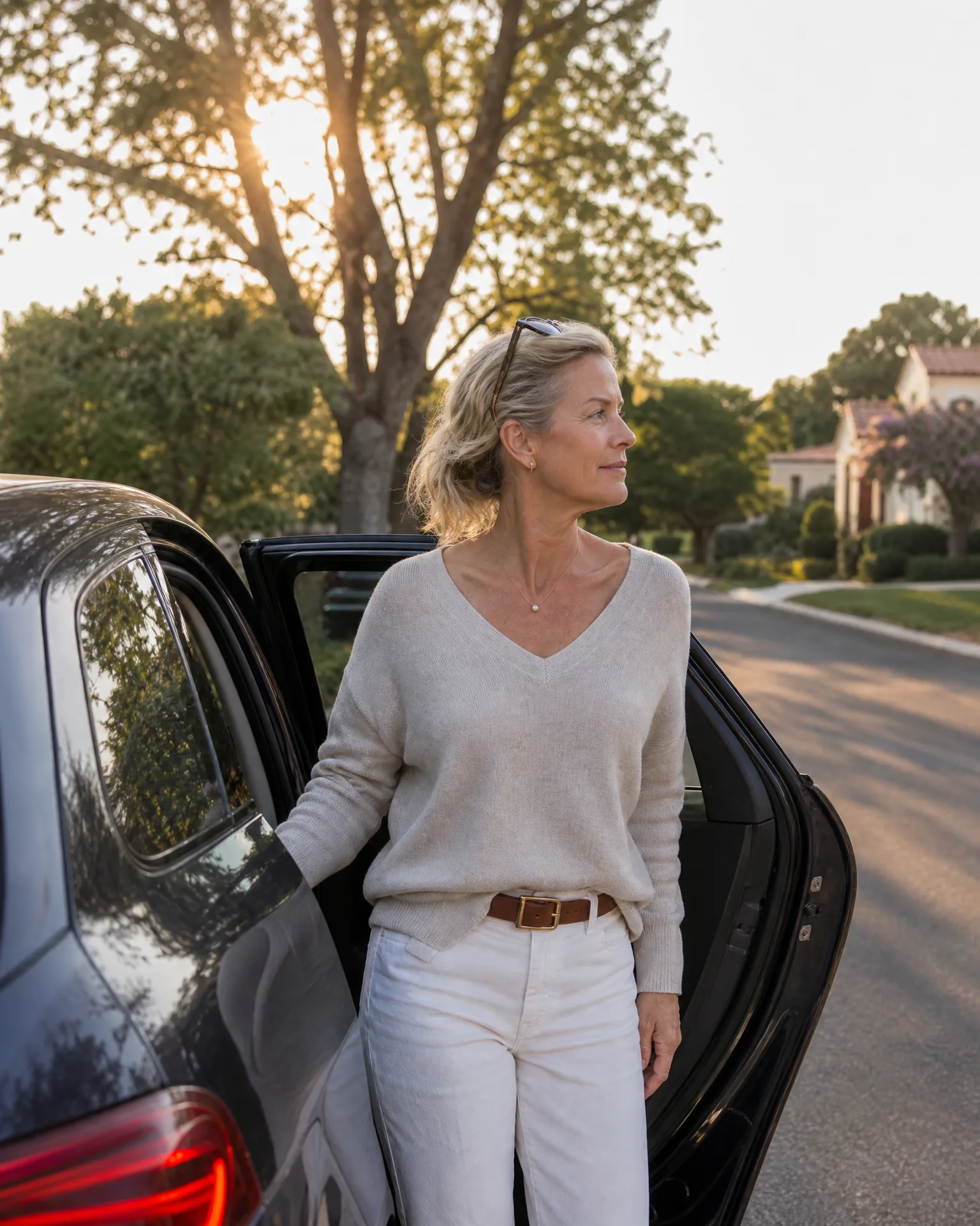 A woman with blonde hair, sunglasses on her head, wearing a light knit sweater, brown belt and white trousers, stands beside the open rear door of a dark car on a residential street with trees backlit by low sun.