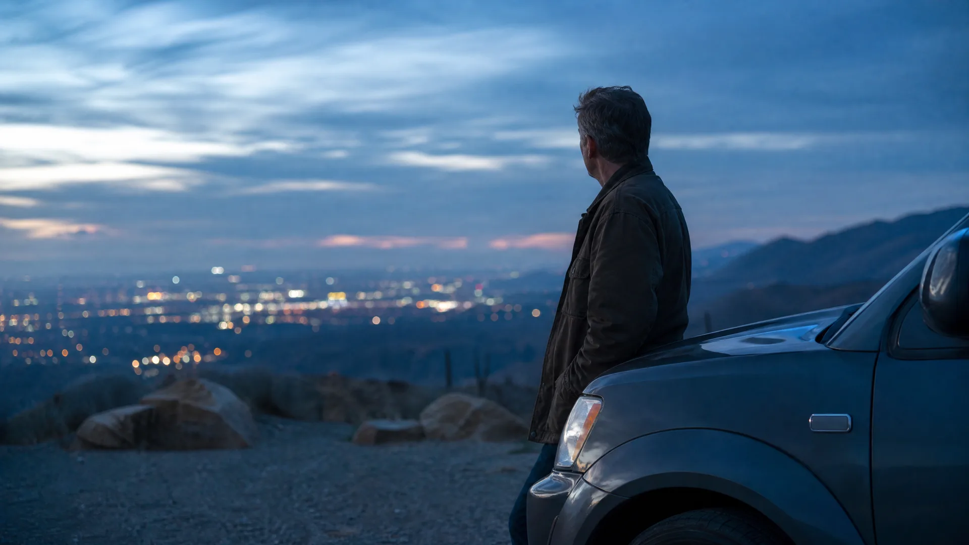 A man in a dark jacket leans against the front of a parked pickup truck on a dirt overlook at dusk, looking out over distant city lights and hills.
