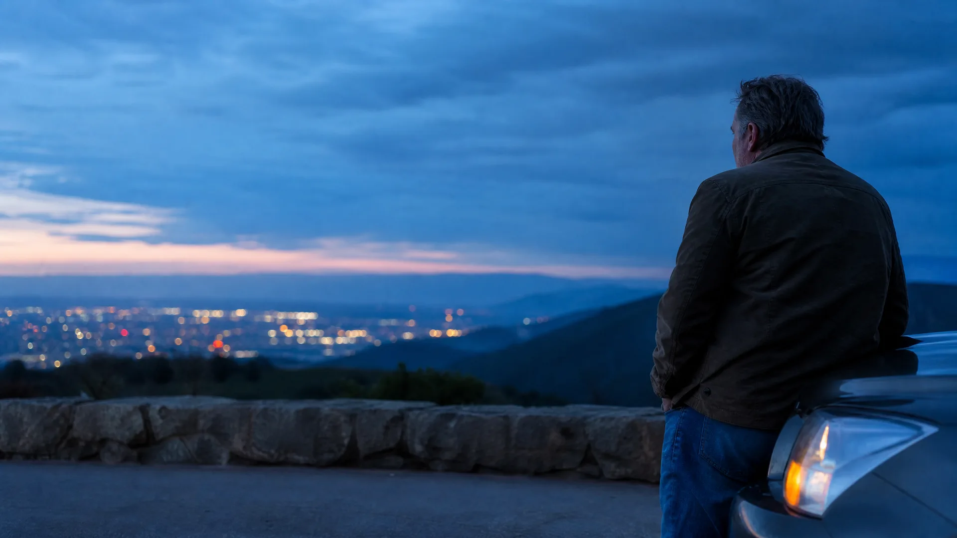 A man in a dark jacket and jeans leans against the front of a car at a stone-walled overlook at dusk, facing away toward a valley of city lights below distant hills.