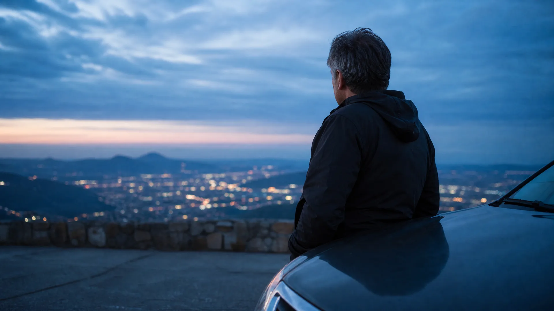A man in a dark hooded jacket leans against the hood of a car at a stone-walled overlook, seen from behind, facing a city of lights in a valley below at dusk.