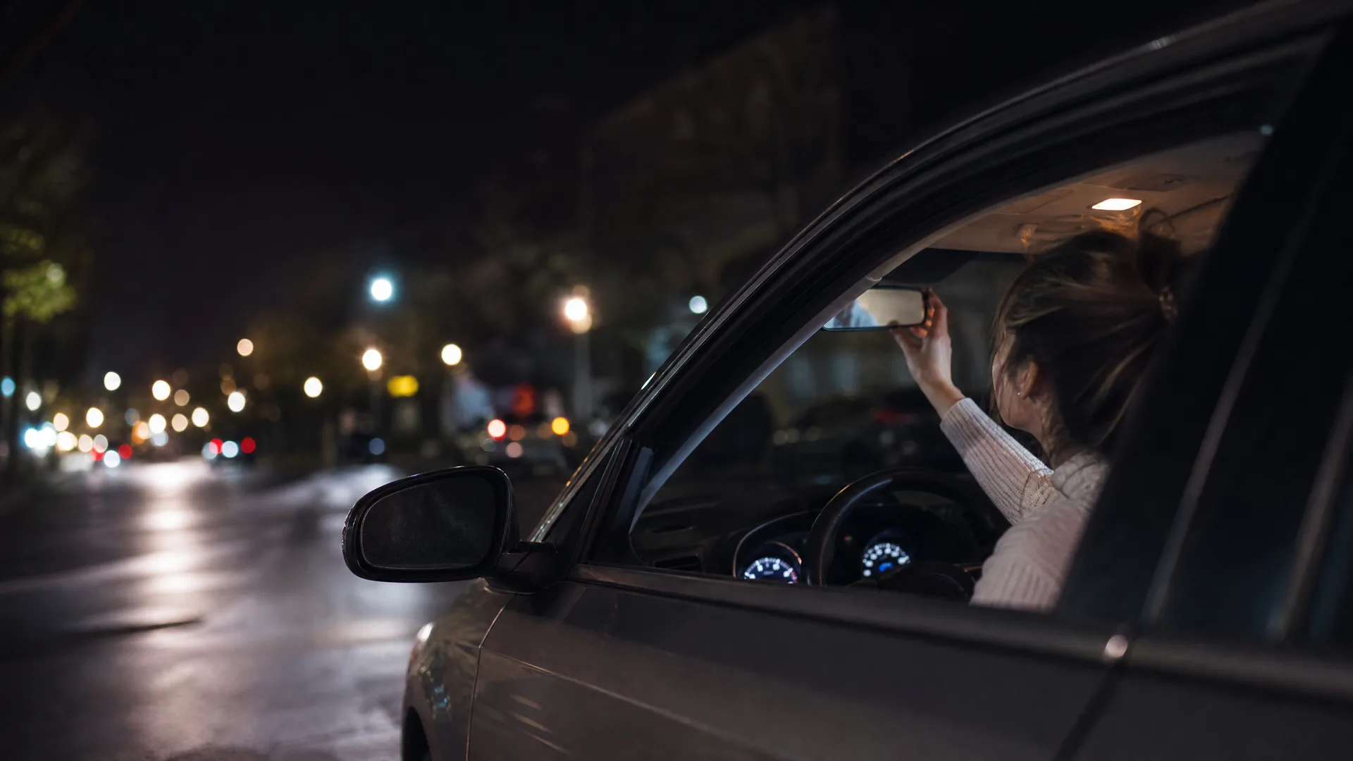 A woman sits in the driver's seat of a car at night, reaching up to adjust the rear-view mirror, seen through the open passenger-side window with blurred street lights behind.