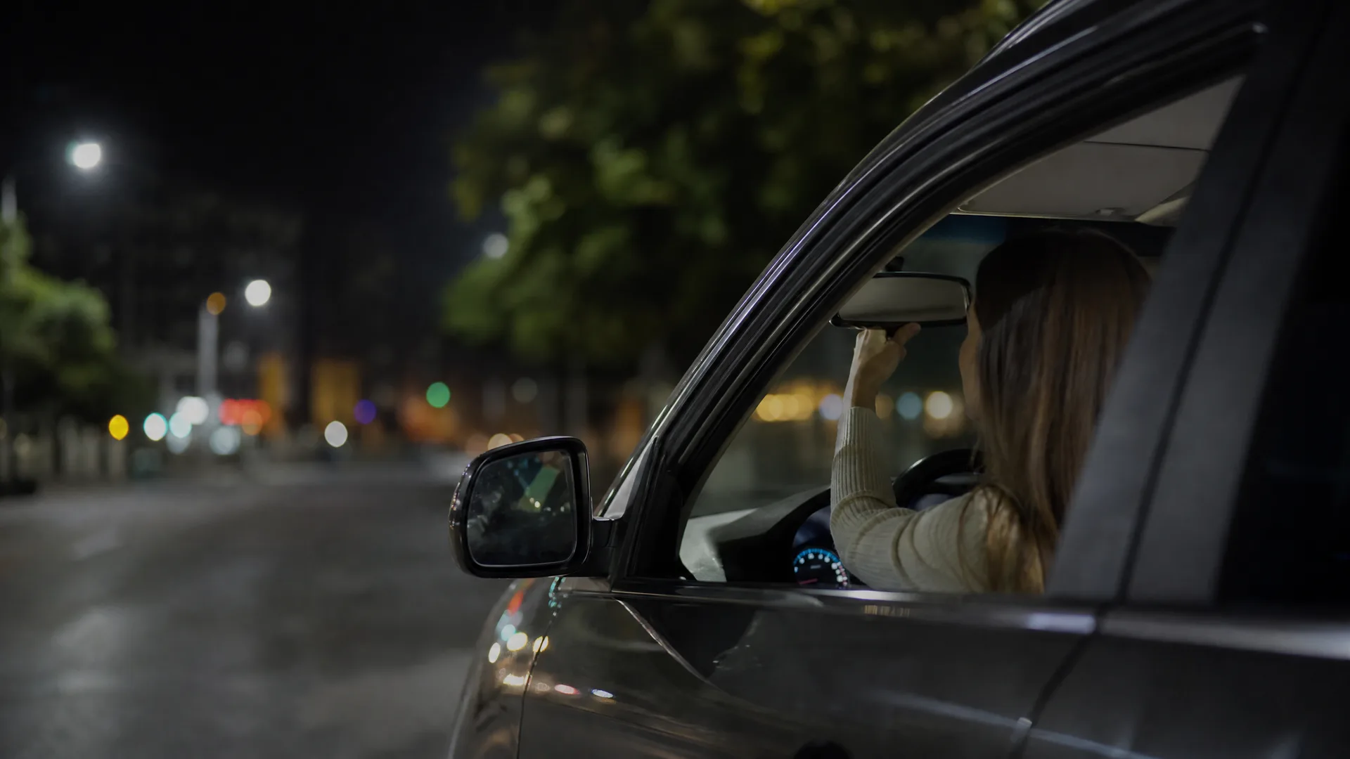 A woman with long hair sits in the driver's seat of a car at night, reaching up to adjust the rear-view mirror, with blurred street lights and trees visible through the open window.